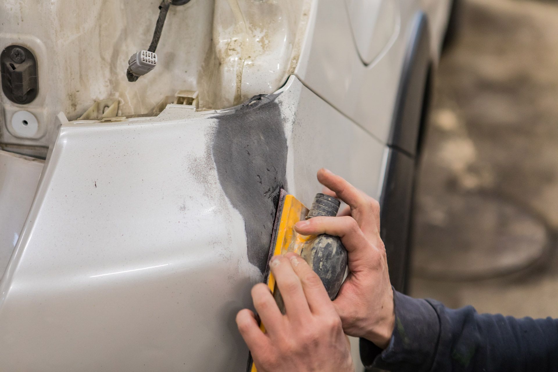 Hands sanding body filler on a damaged car panel, near the rear tire.
