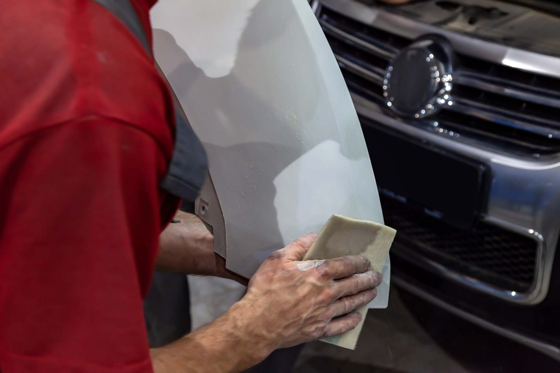 Mechanic sanding a car panel with body filler, near a vehicle's front bumper.