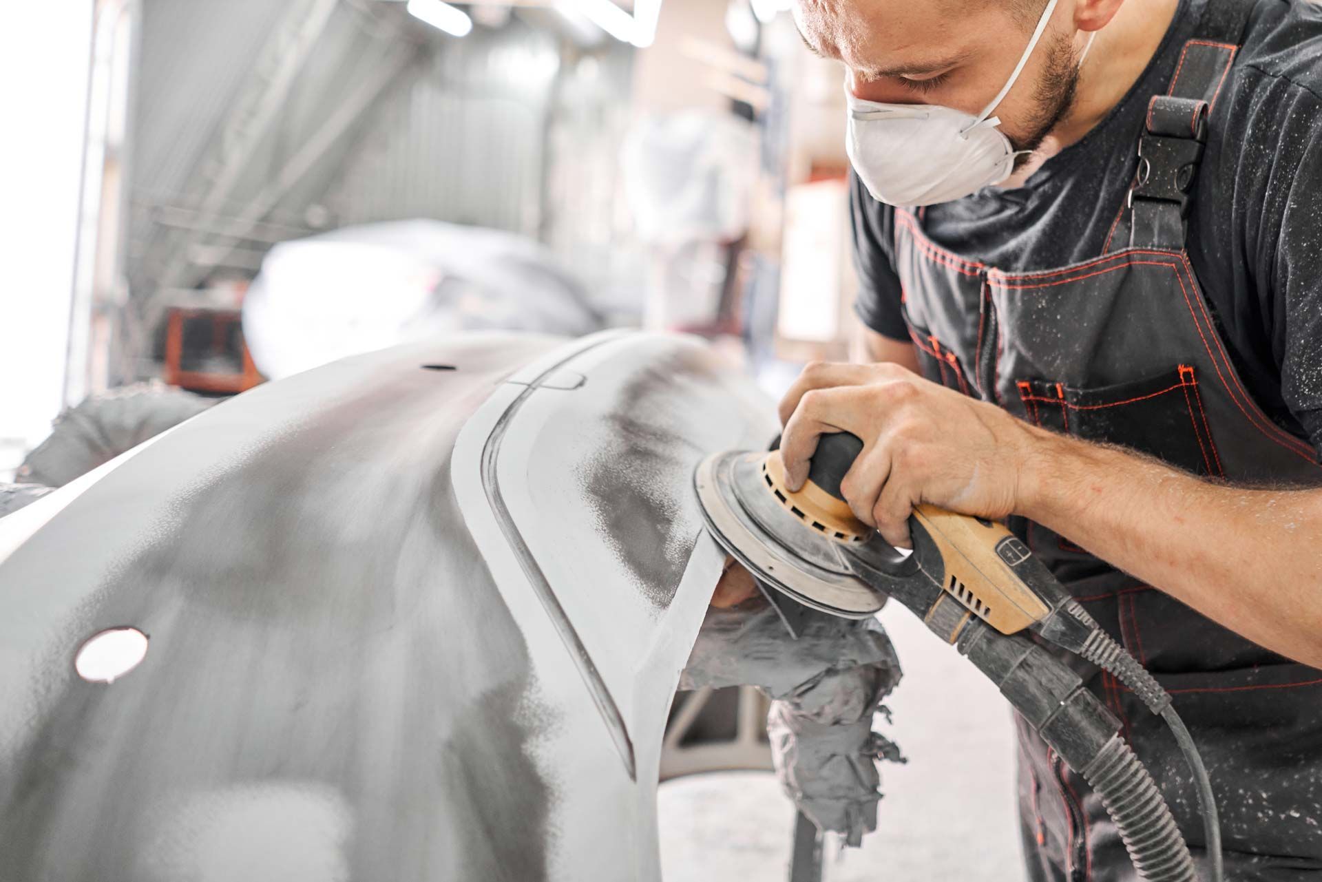 Person with respirator sanding car bumper in a workshop.