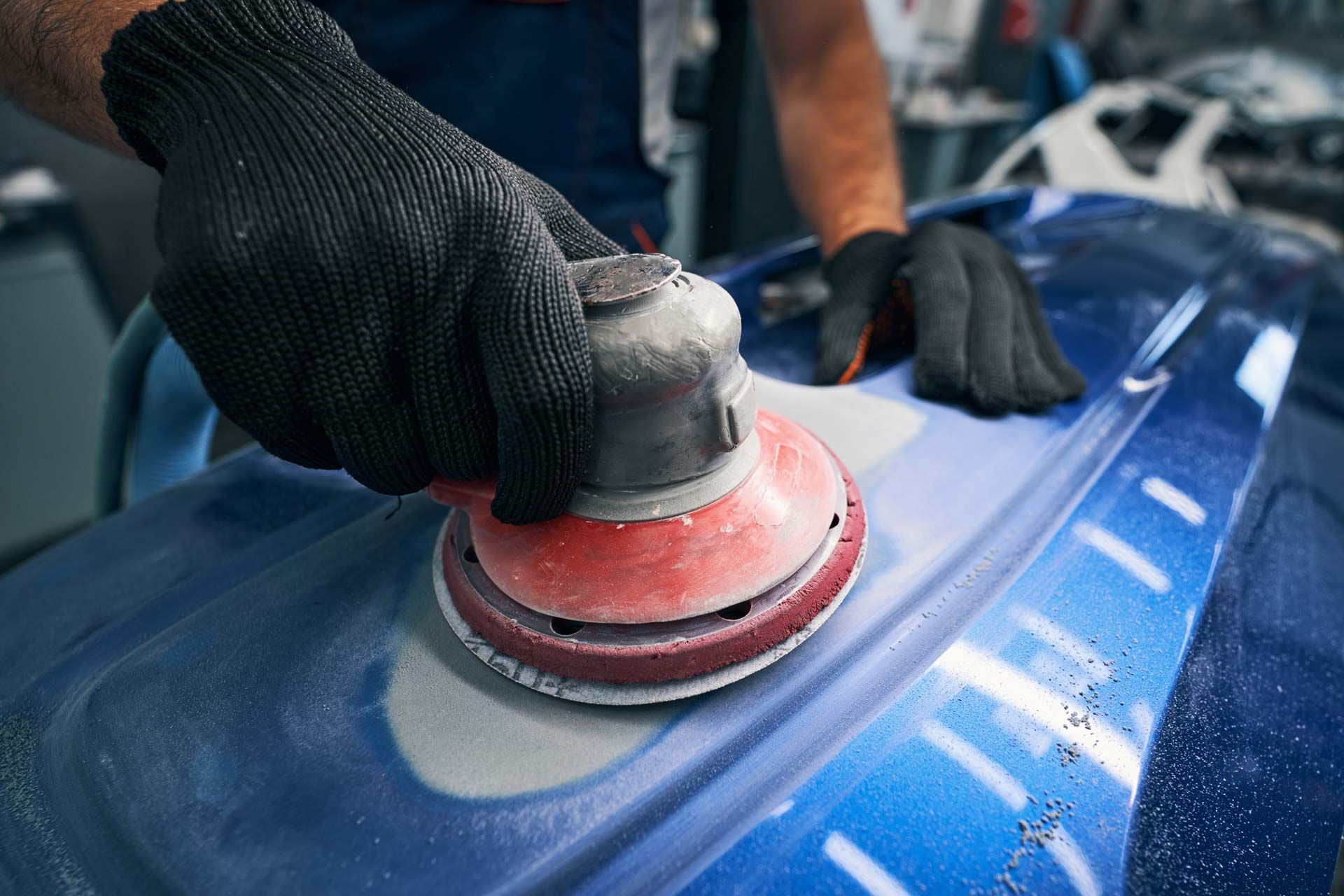 Person sanding blue car part with a red and gray power sander, wearing black gloves.