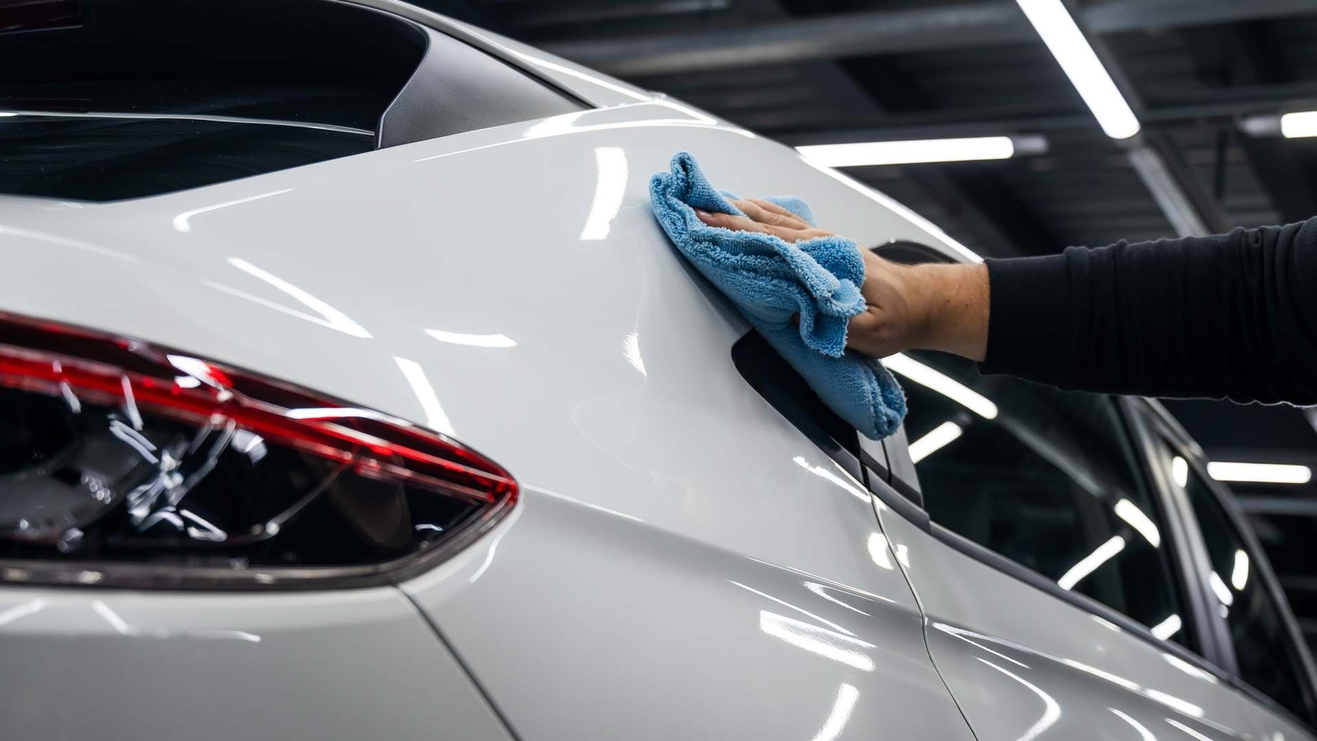 Person's hand wiping a white car with a blue microfiber cloth, in a well-lit garage.