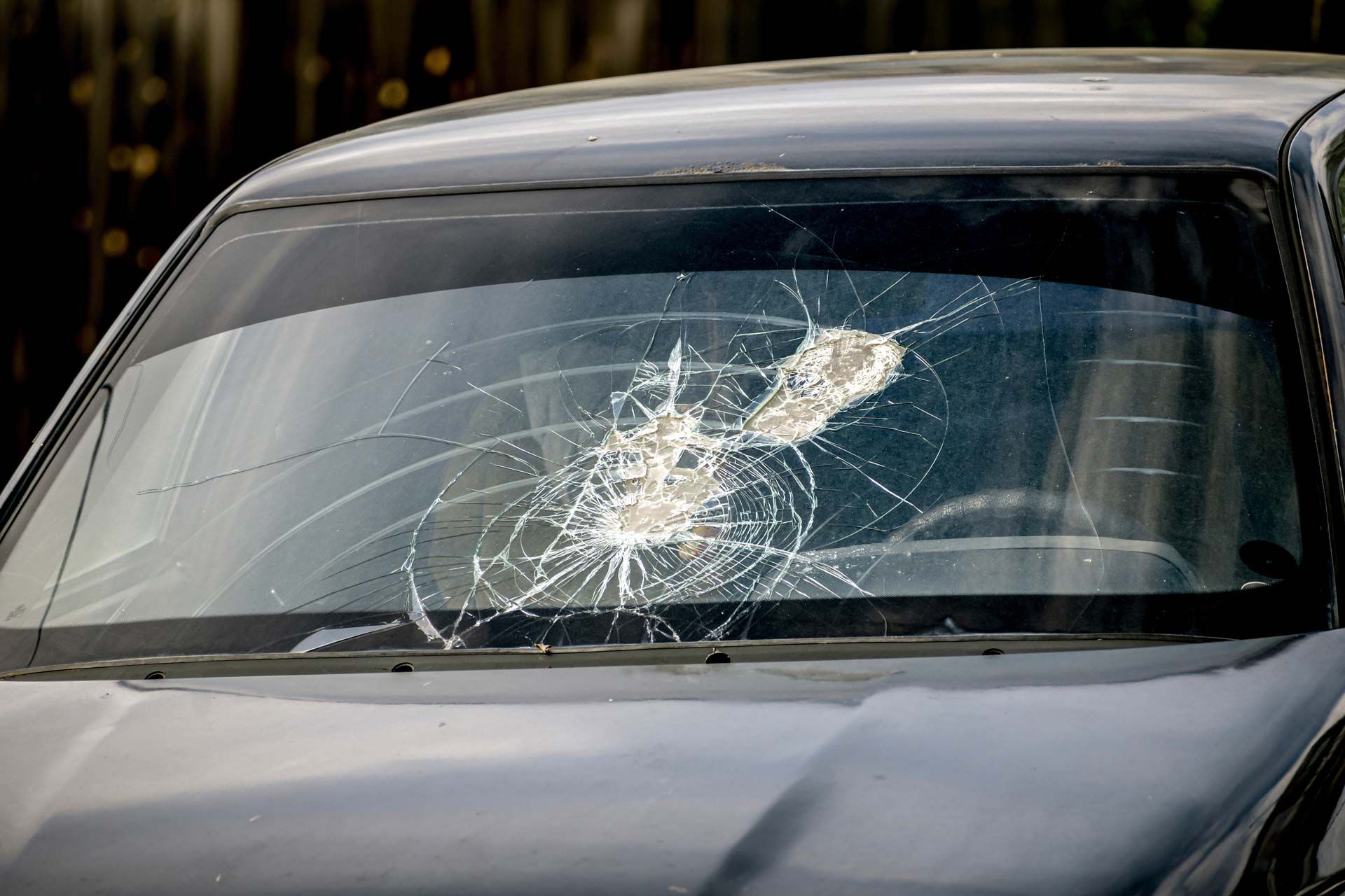 Cracked car windshield showing a central impact point with radiating fractures, likely due to a collision.