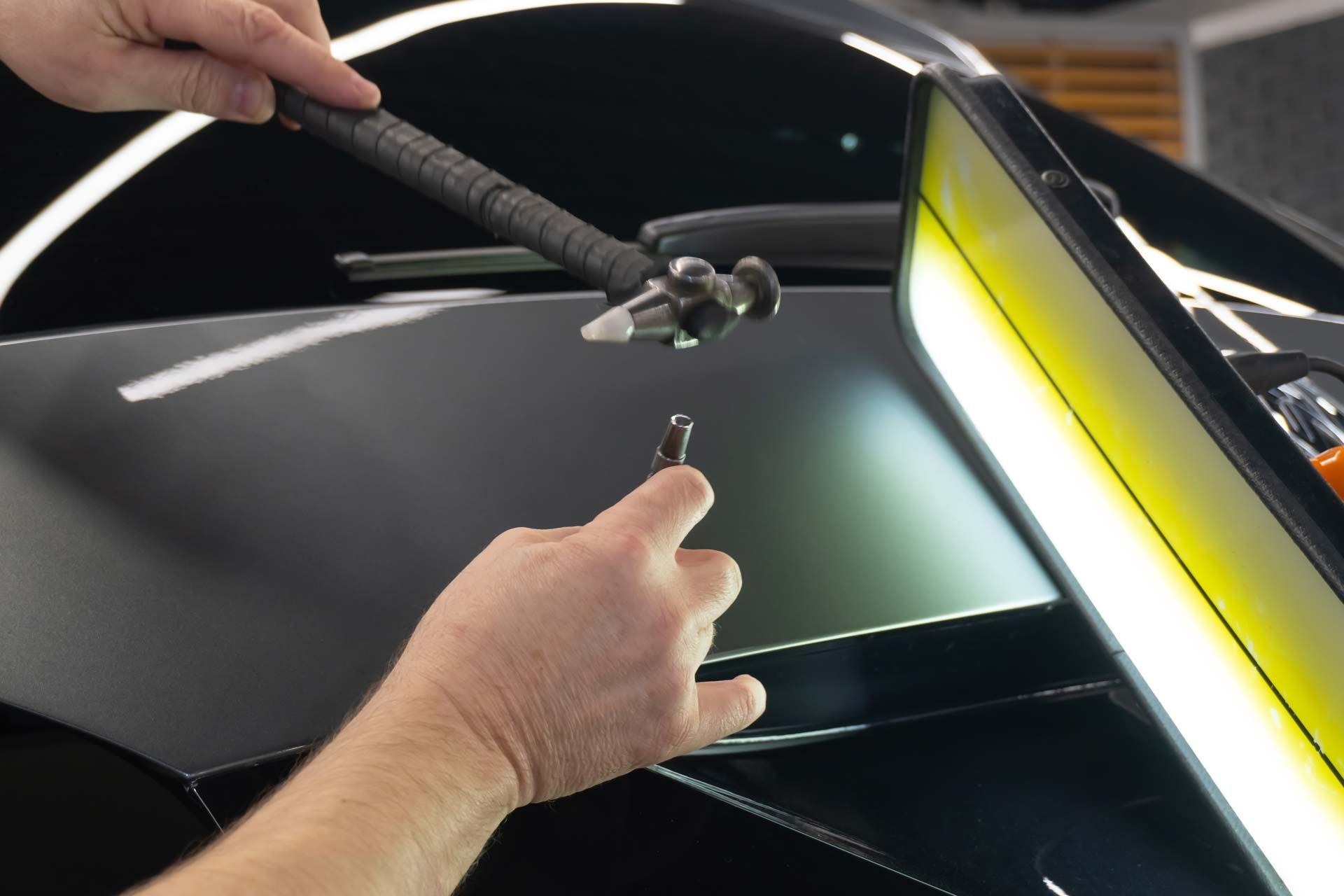 Hands using tools to remove a dent from a black car panel.