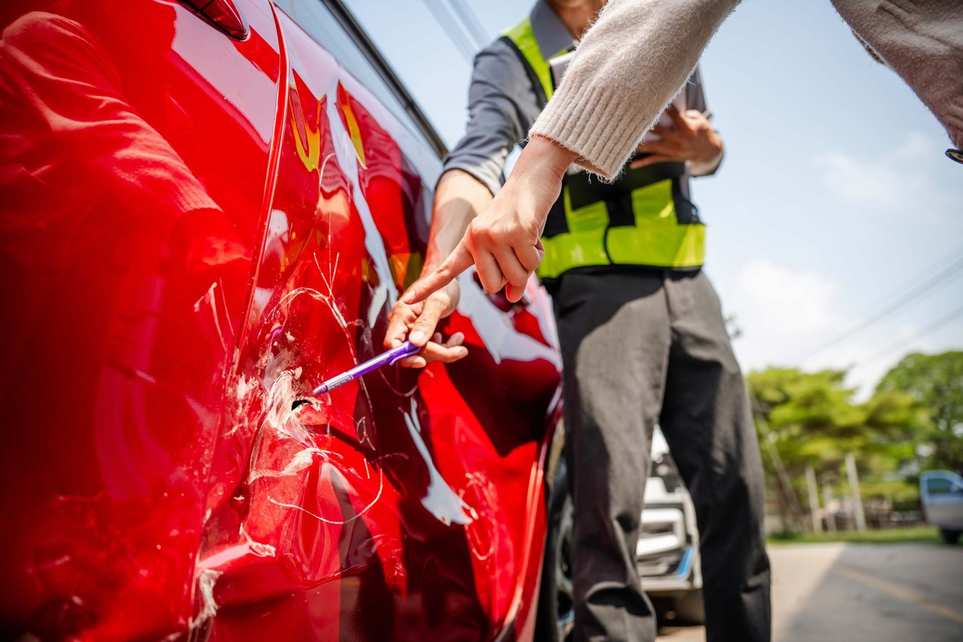 Person points at damage on a red car with an insurance adjuster, outdoors.