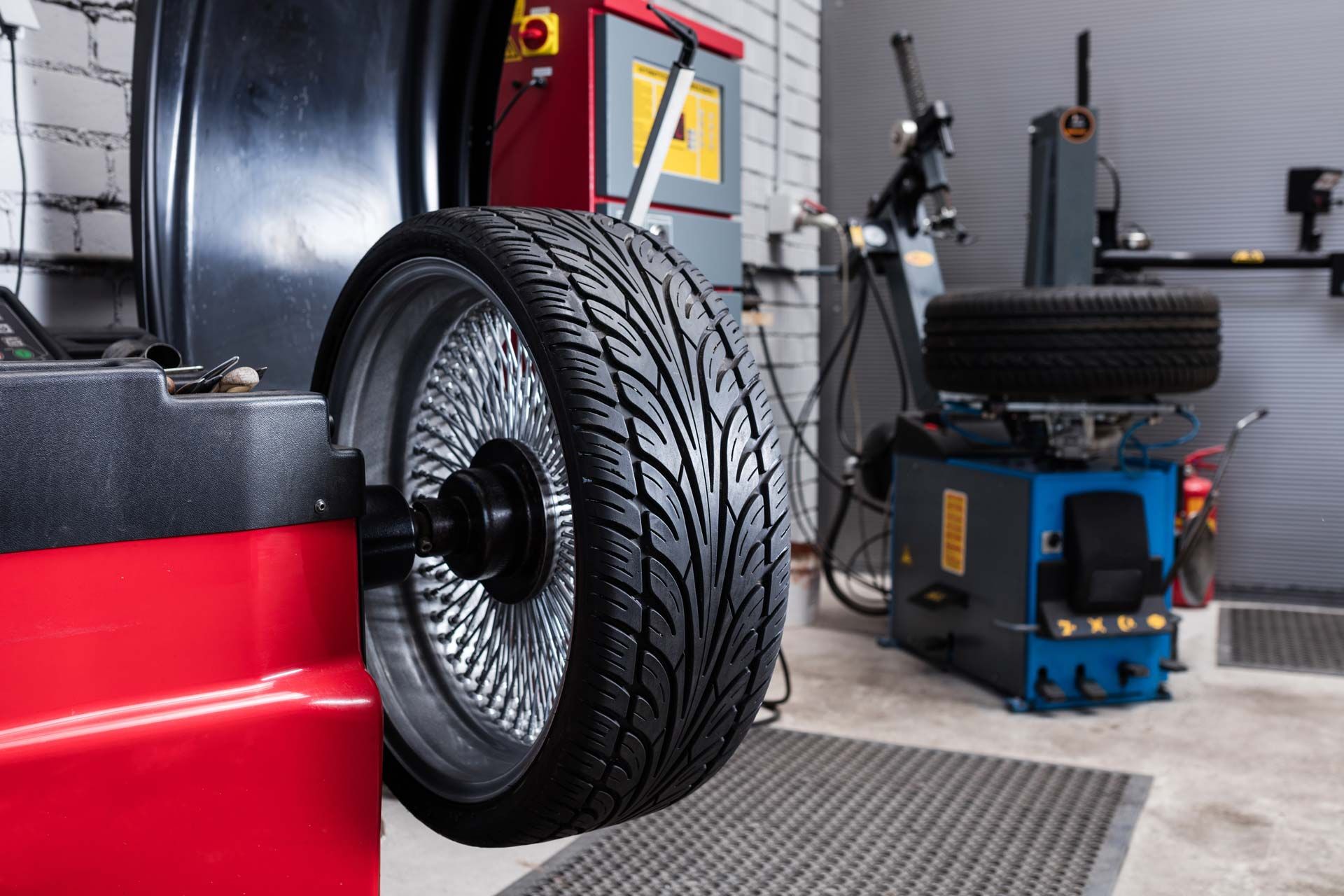 Tire on a balancing machine in a garage; red machine, dark tire, silver rim, blue machine in background.