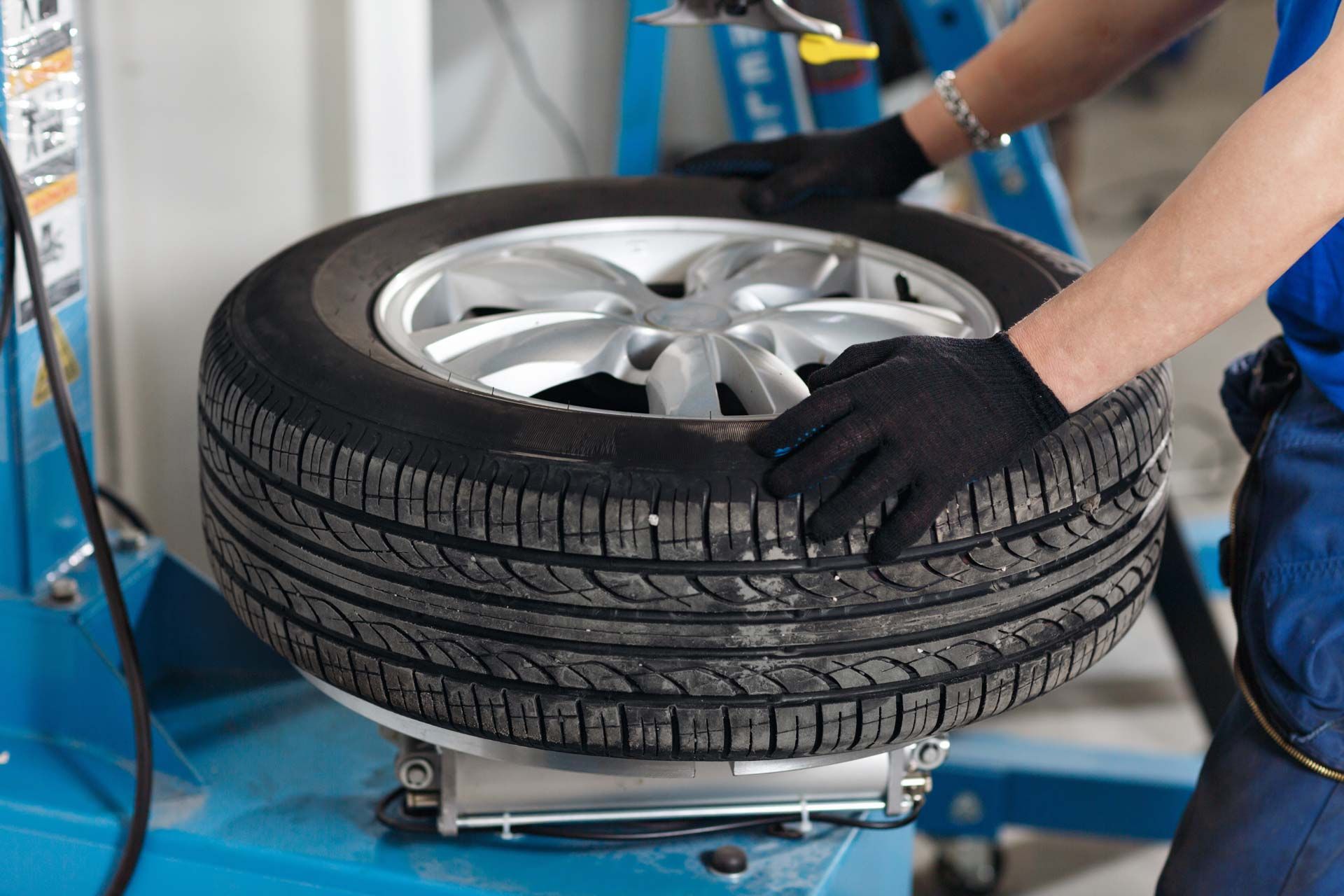 Mechanic working on a tire using a machine; wearing black gloves, blue coveralls.