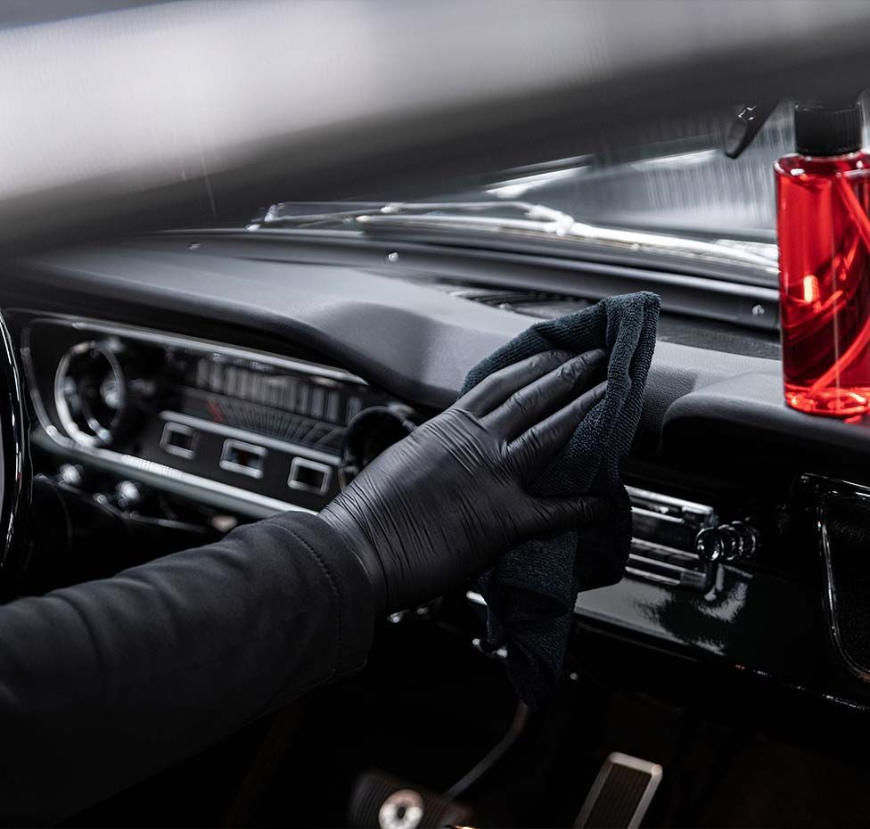 A person wearing black gloves wiping a car's black dashboard with a cloth, cleaning spray bottle on the right.