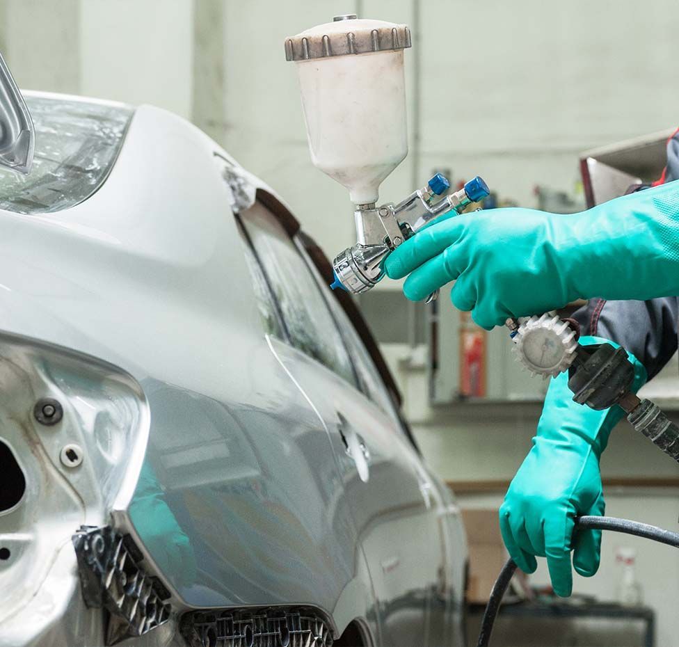 A person in teal gloves sprays paint on a car's damaged body.
