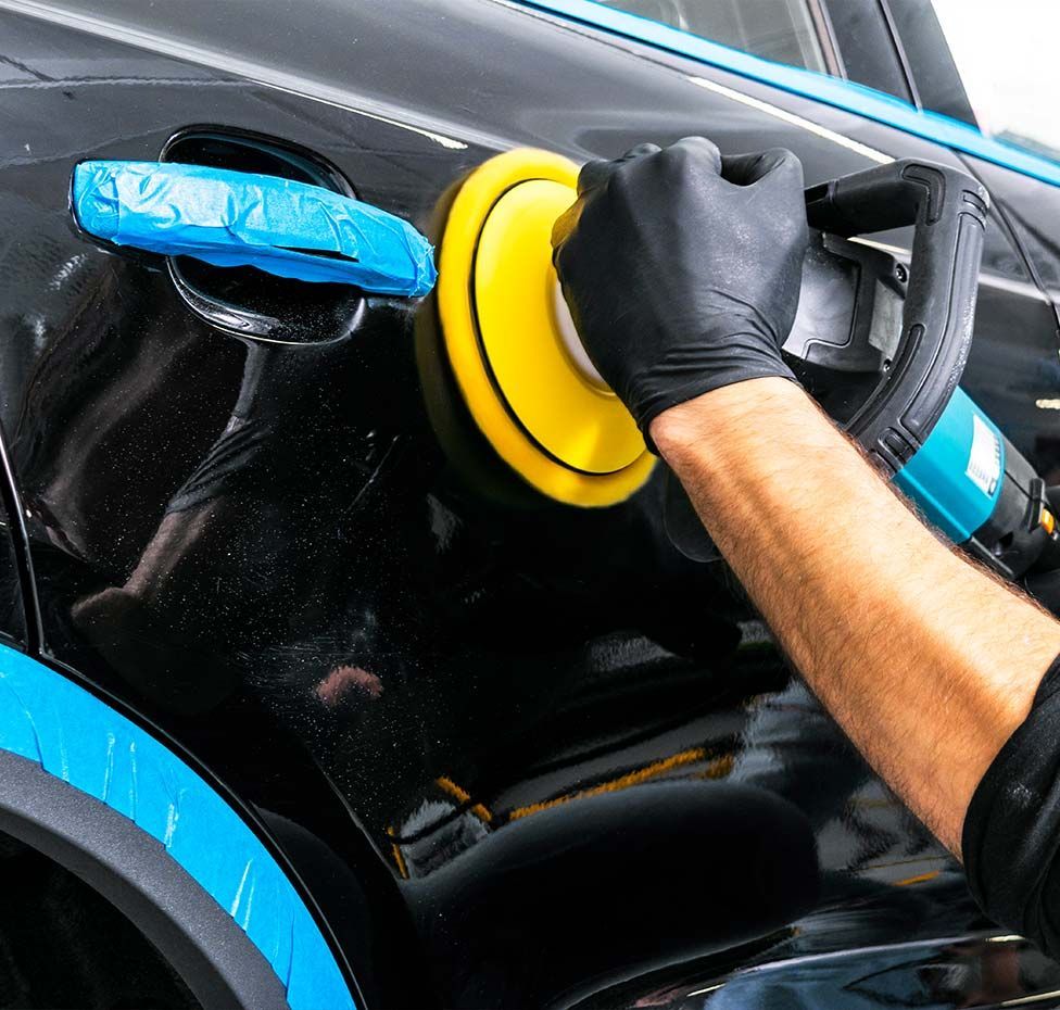 Person using a power buffer to polish a black car's paint, wearing black gloves.
