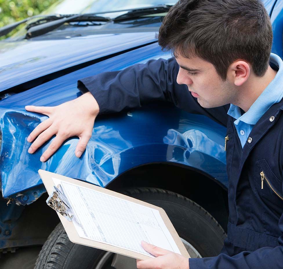 Person inspecting damaged blue car, holding clipboard.