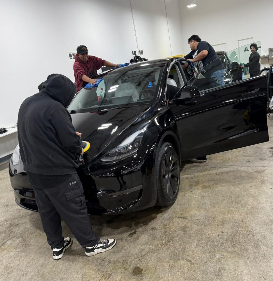 Three people cleaning a black Tesla car inside a brightly lit garage.