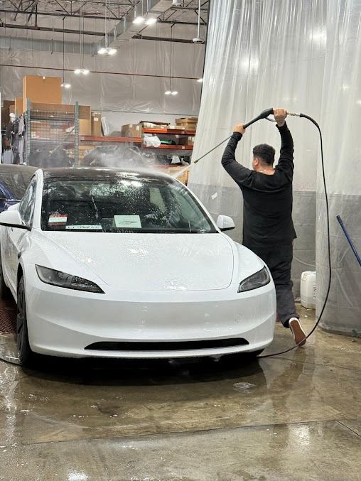 A person washing a white Tesla car with a hose in a warehouse setting.