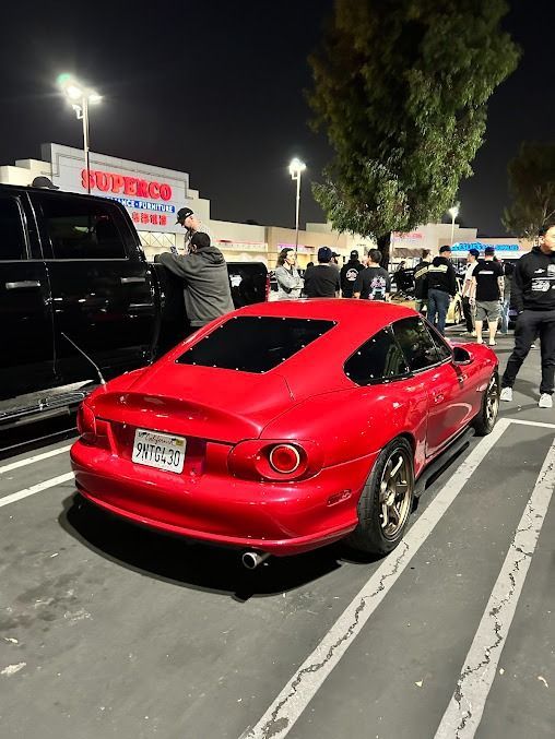 Red Mazda Miata with a hardtop parked in a crowded parking lot at night.