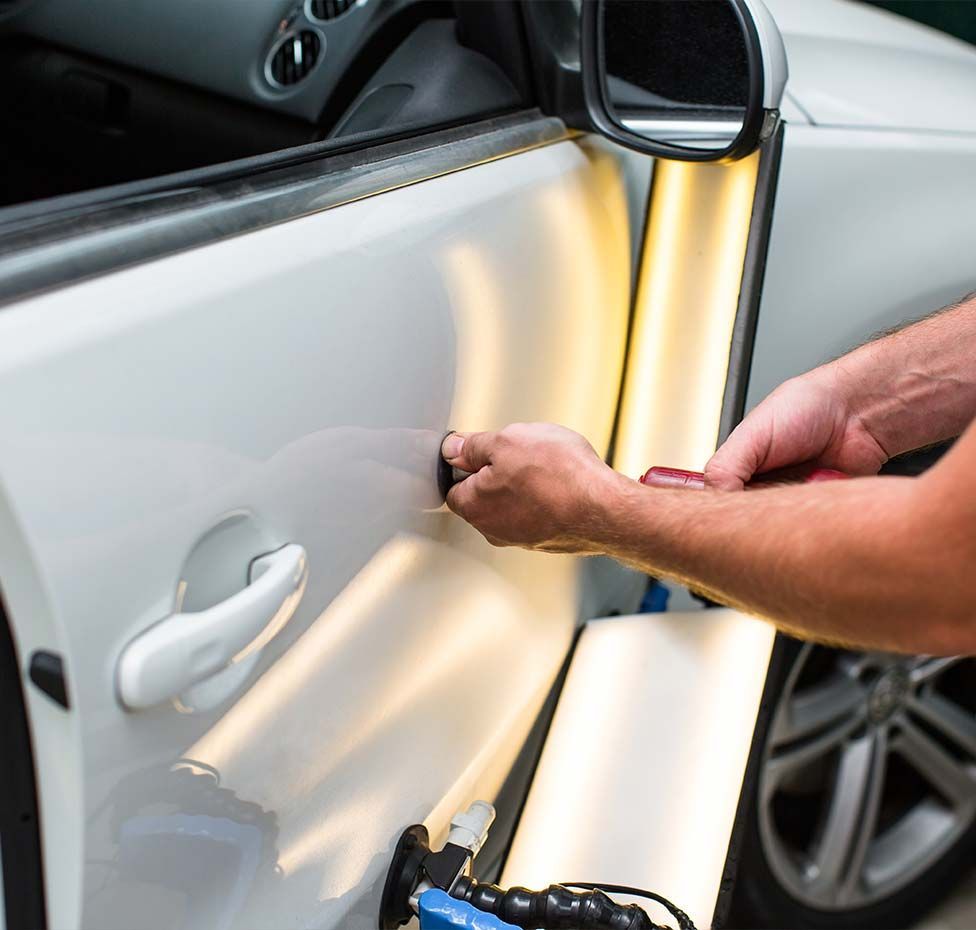 Person using a tool to repair a dent on a white car door, illuminated by a light.