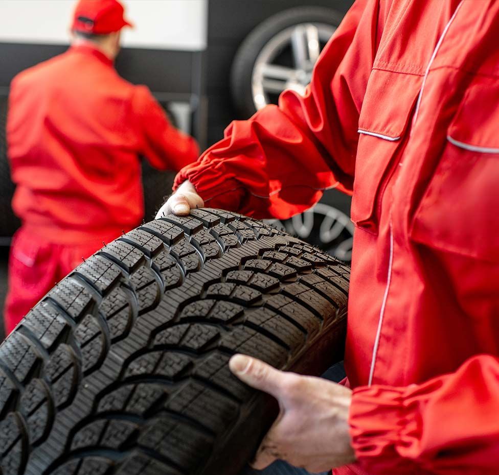 Mechanic in red overalls carrying a tire in a garage, another mechanic in the background.