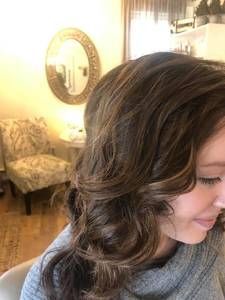 Woman with wavy brown hair, looking right, in a well-lit room with a decorative mirror and chair
