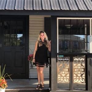 Woman in black dress stands in doorway of building with black door, windows, and decorative metalwork