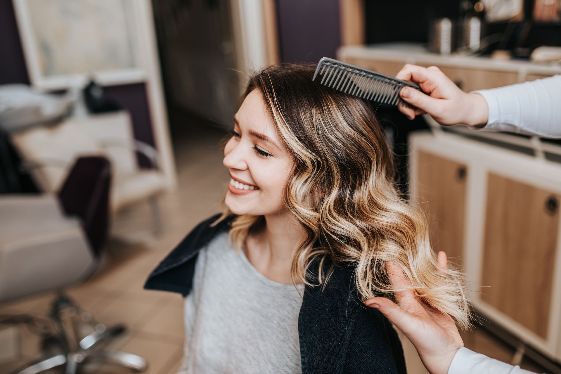 Woman smiling while a person combs her highlighted, wavy hair in a salon