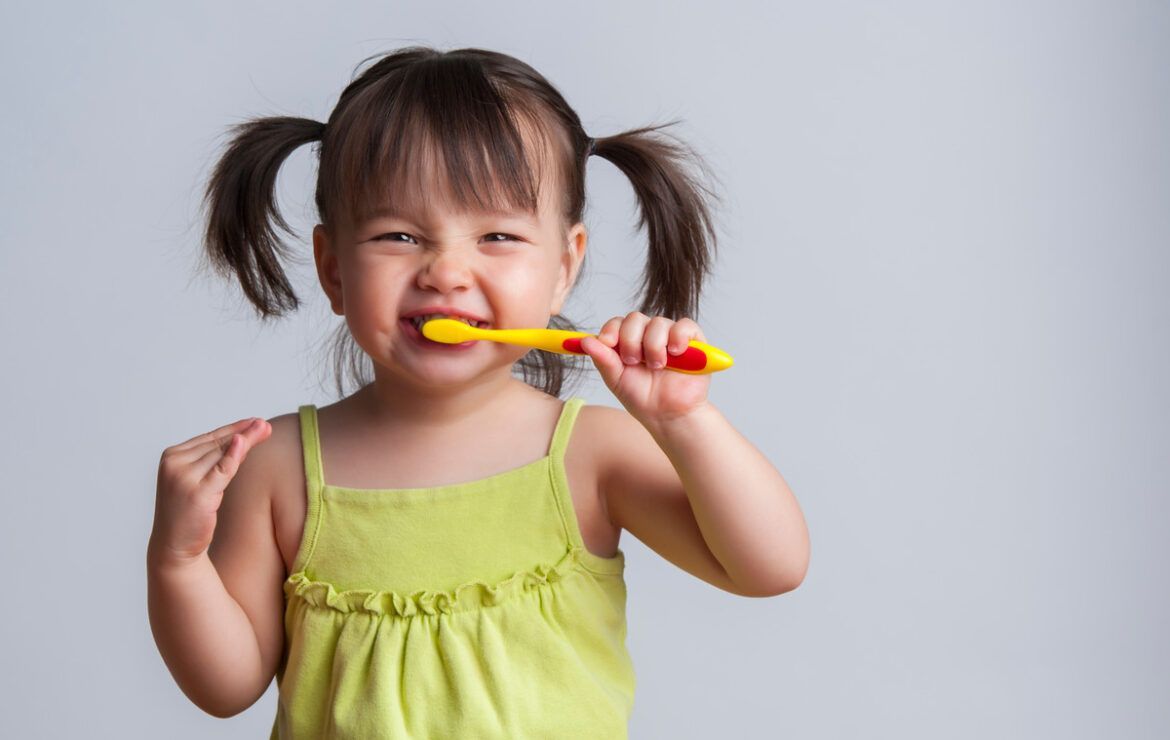 A little girl is brushing her teeth with a yellow toothbrush.