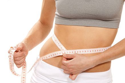 Mujer midiendo su cintura con una cinta métrica blanca, uvestida con un top gris y pantalones blancos.