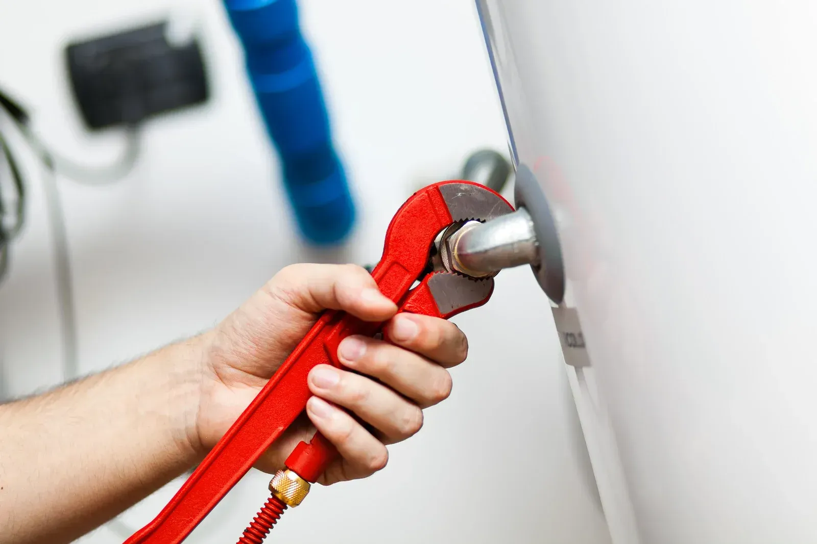 A hand holding a red pipe wrench, tightening a metal pipe on a white appliance.