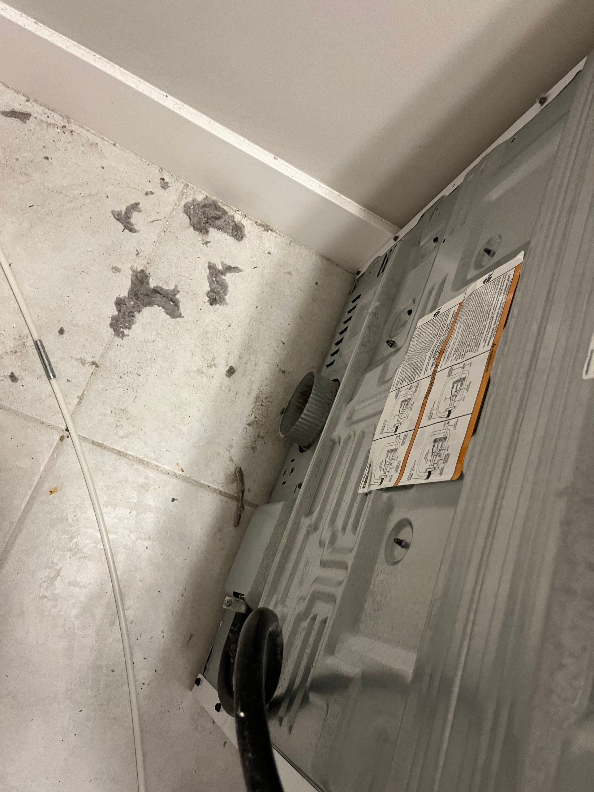 A close-up of a washing machine's back panel in a corner. The floor and wall are light-colored, with lint and debris.