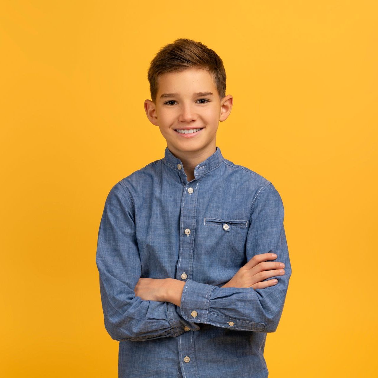 A young boy in a denim shirt is standing with his arms crossed on a yellow background.