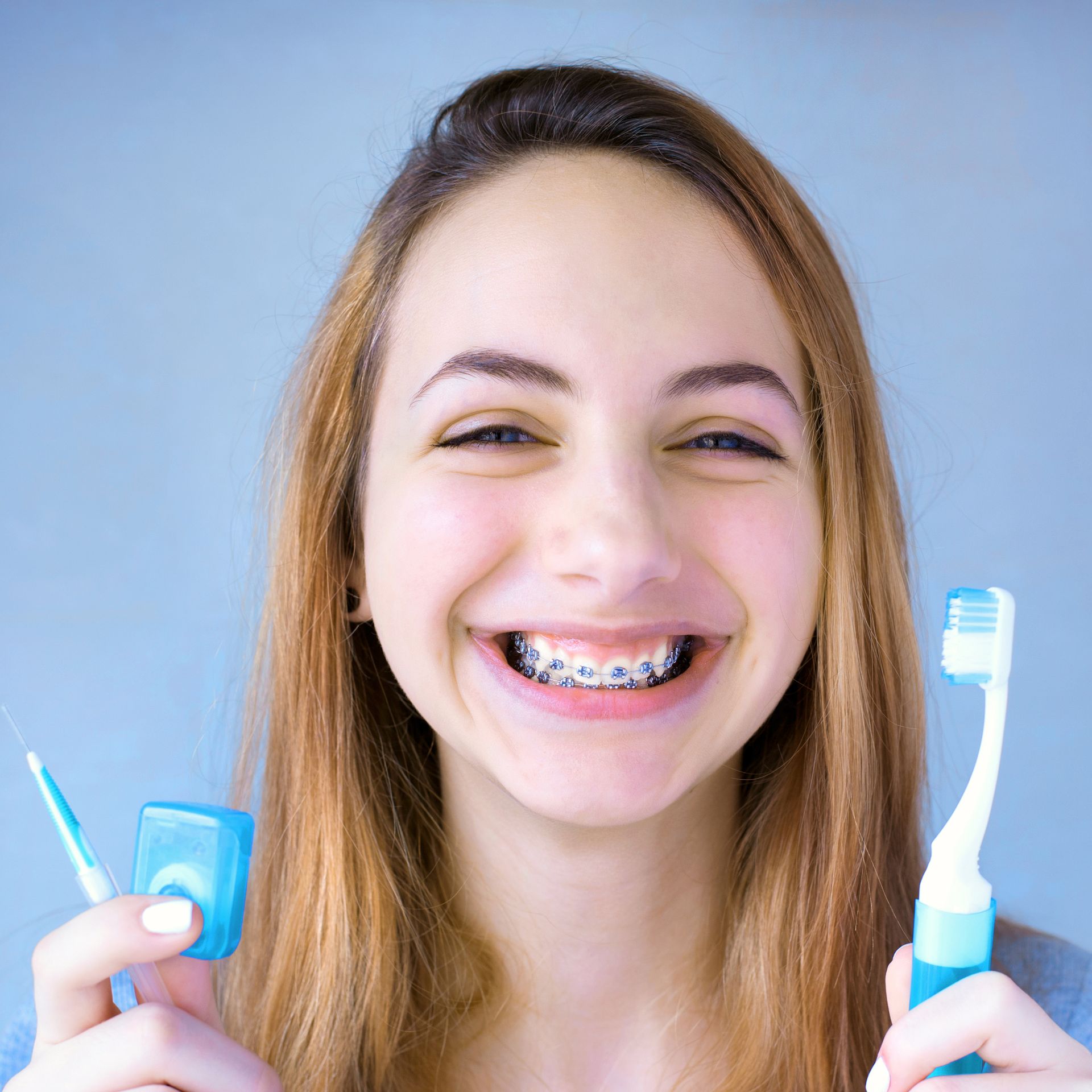 A woman with braces is smiling while holding a toothbrush and floss