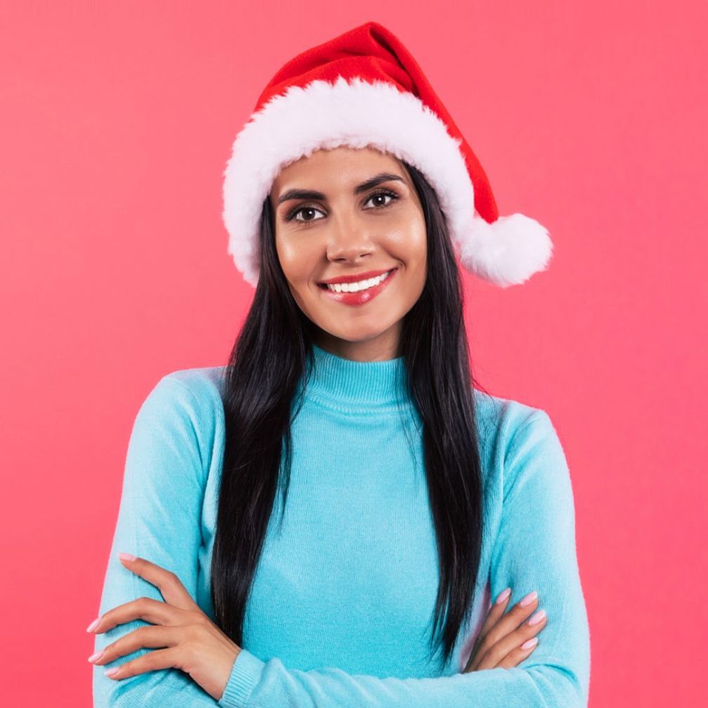 A woman wearing a santa hat and a blue sweater is smiling with her arms crossed.