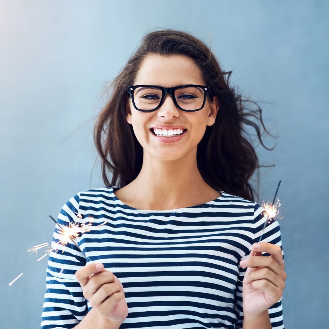 A woman wearing glasses and a striped shirt is holding sparklers and smiling.