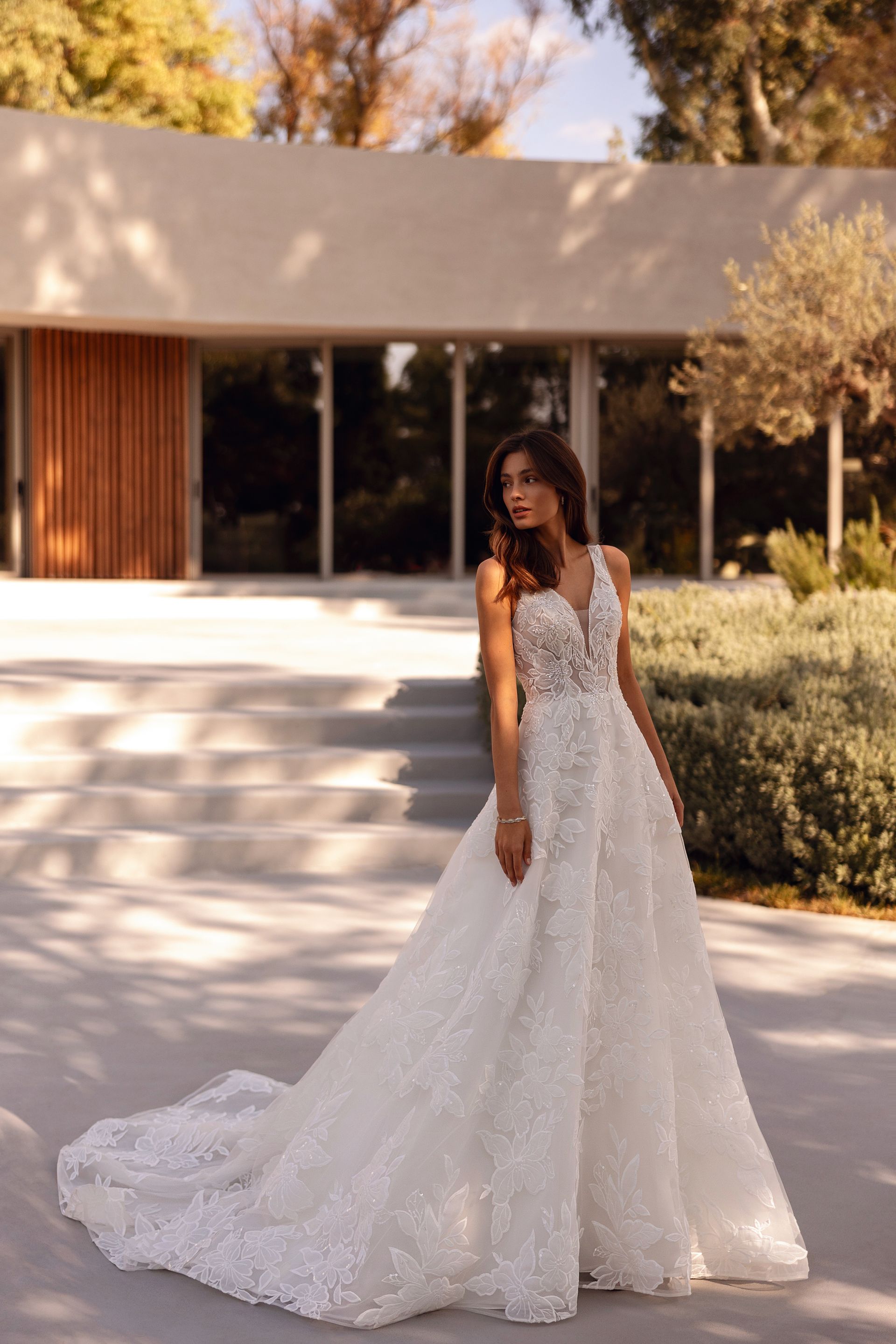 Donna in abito da sposa bianco con strascico, in piedi sui gradini di fronte a un edificio moderno.