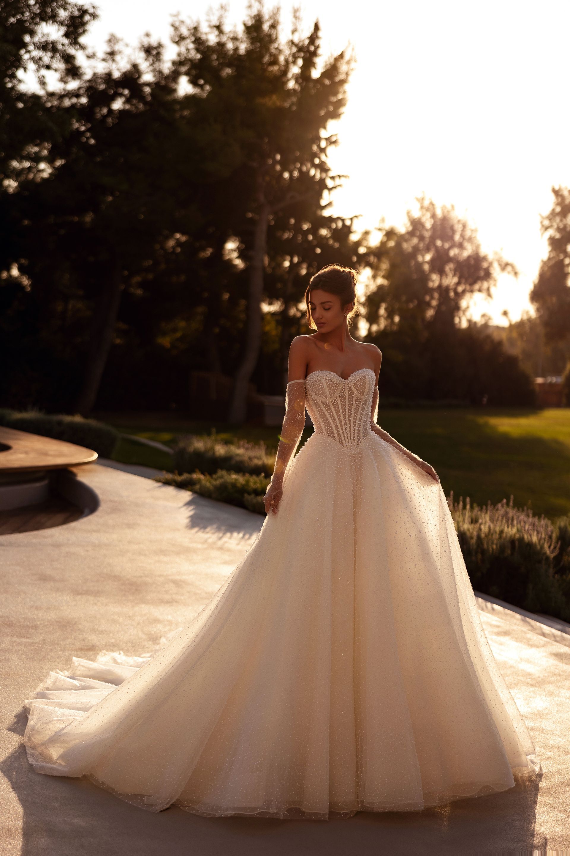 Una donna con un abito da sposa color avorio senza spalline è in piedi su un patio, la luce del sole illumina la scena.