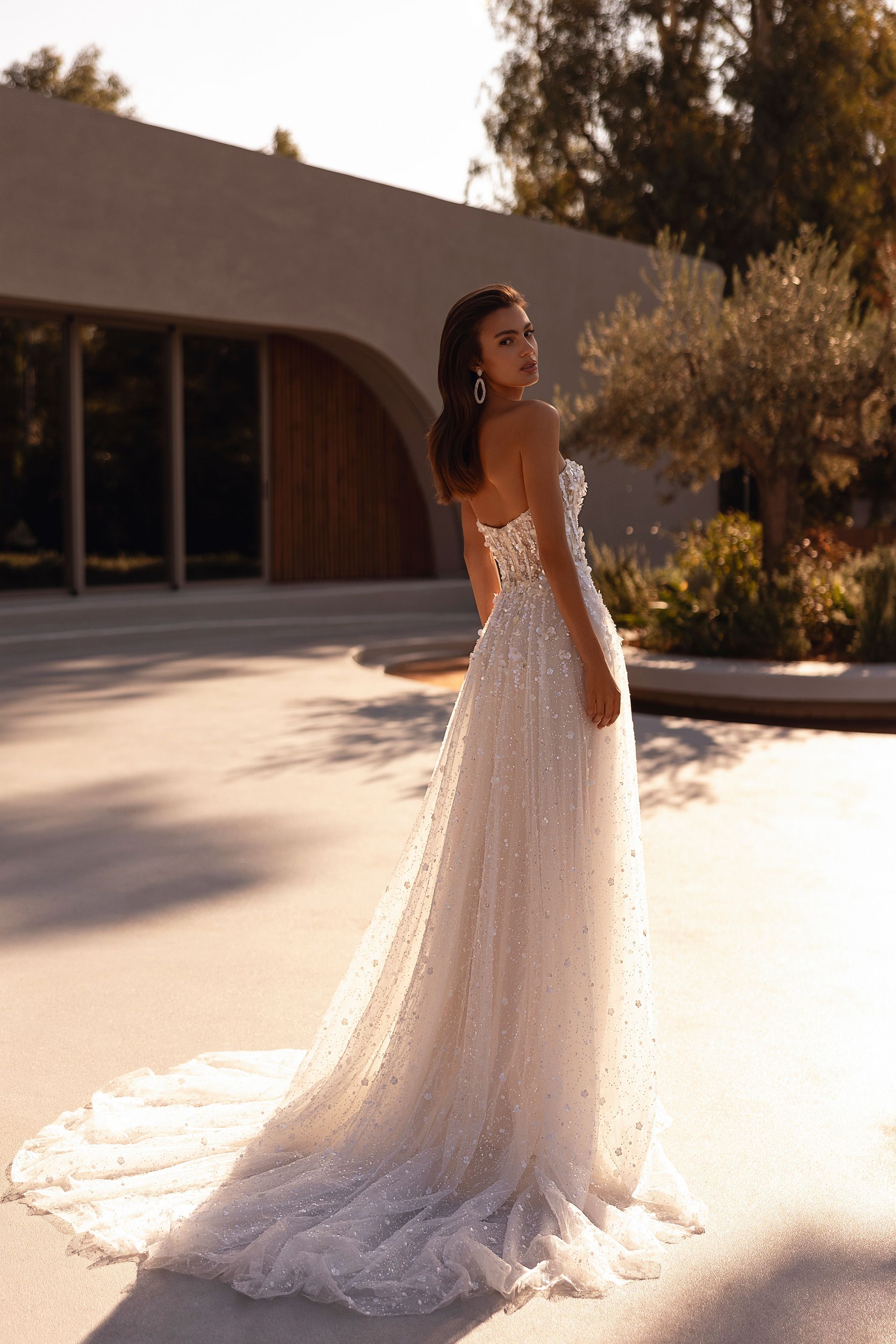 Donna in abito da sposa senza spalline con perline e lungo strascico, in piedi fuori da un edificio moderno.