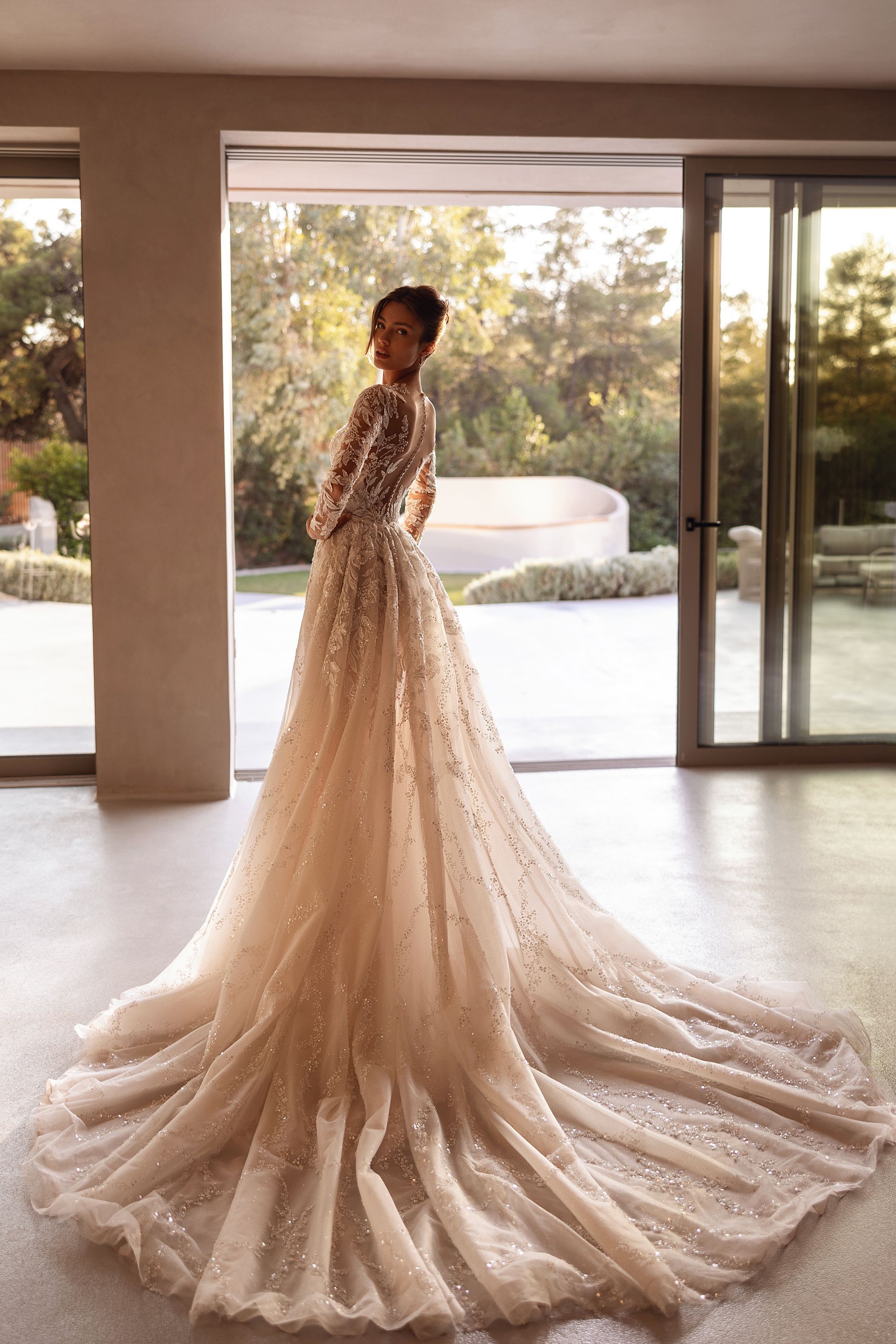 Donna con un abito scintillante e fluente, con un lungo strascico, in piedi vicino a grandi finestre in un interno luminoso.