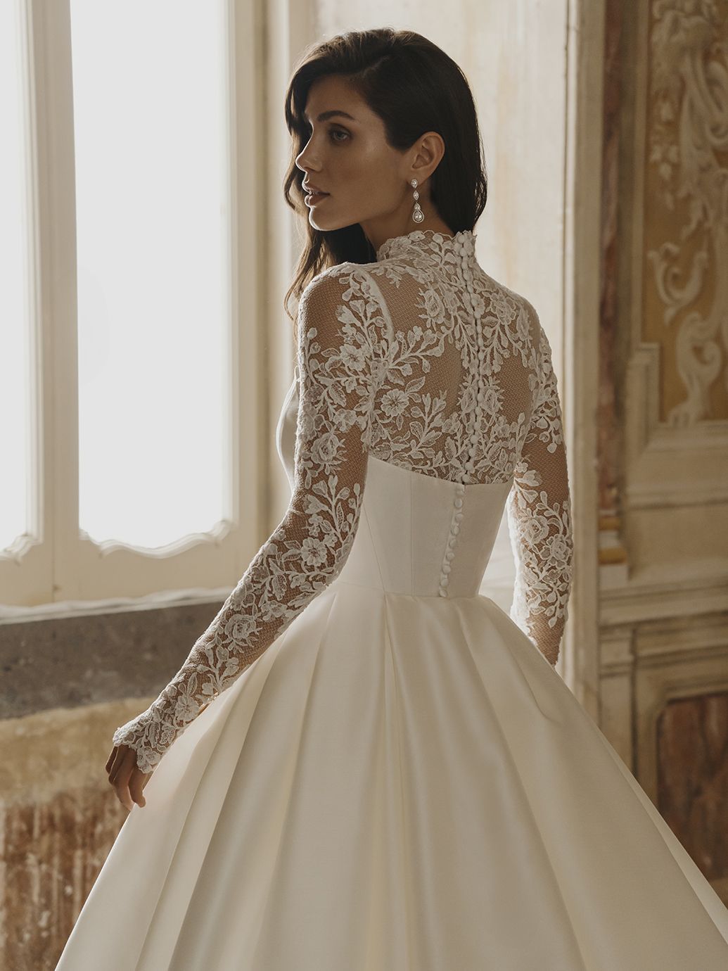 Donna in abito da sposa in raso bianco con maniche in pizzo e scollatura alta, in piedi vicino a una finestra.