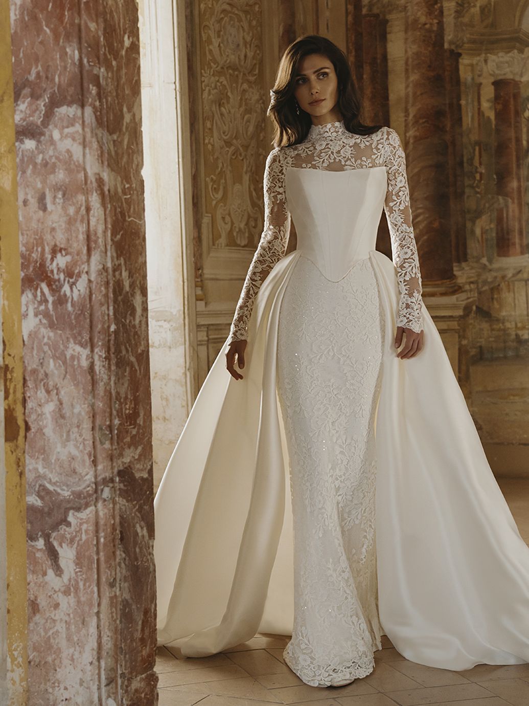 Donna in abito da sposa bianco con lunghe maniche in pizzo e strascico fluente, in piedi in una grande sala.