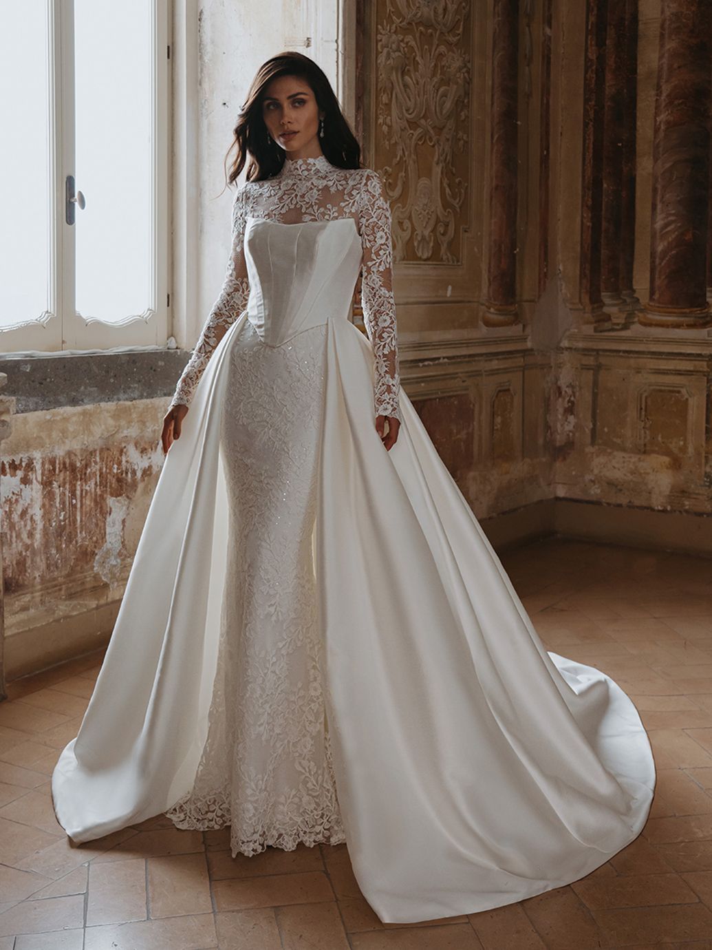 Donna in abito da sposa bianco con maniche in pizzo e lungo strascico, in piedi in un interno.