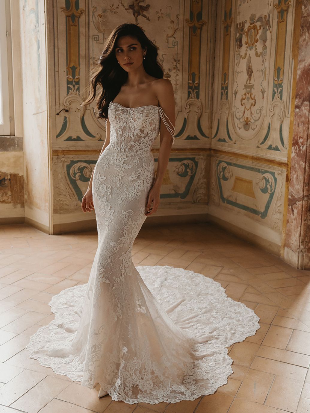 Donna con abito da sposa a sirena in pizzo bianco senza spalline, strascico sul pavimento; in piedi in una stanza.