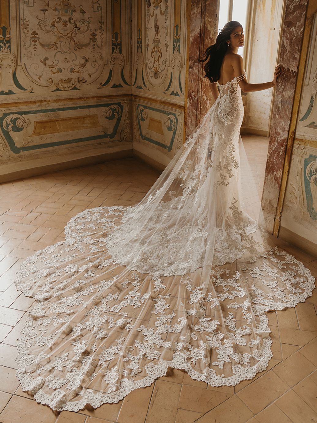 Donna in abito da sposa in pizzo con lungo strascico, in piedi vicino a una finestra in una stanza con dipinti.