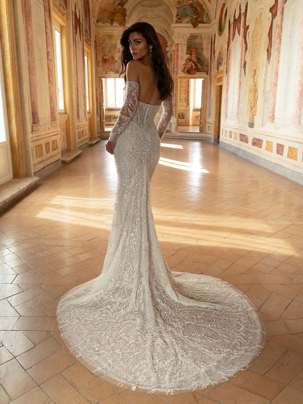 Donna in un abito scintillante con spalle scoperte e lungo strascico, in piedi in un corridoio decorato con pareti dipinte.