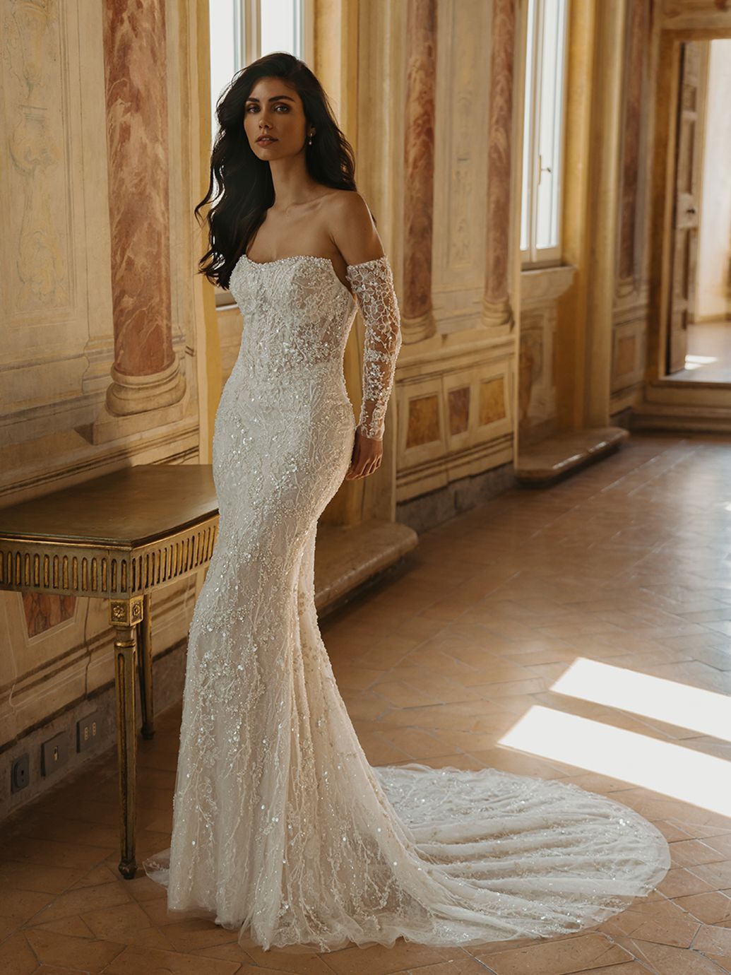 Donna in abito a sirena senza spalline con perline, lunghe maniche trasparenti e strascico. 