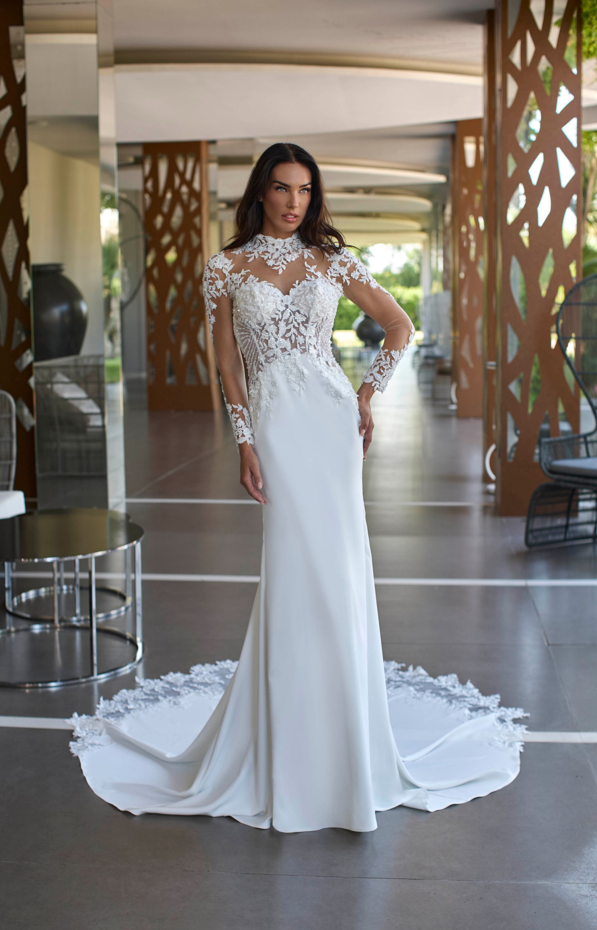Donna in abito da sposa bianco con corpetto in pizzo, maniche lunghe e strascico, in piedi in un interno.