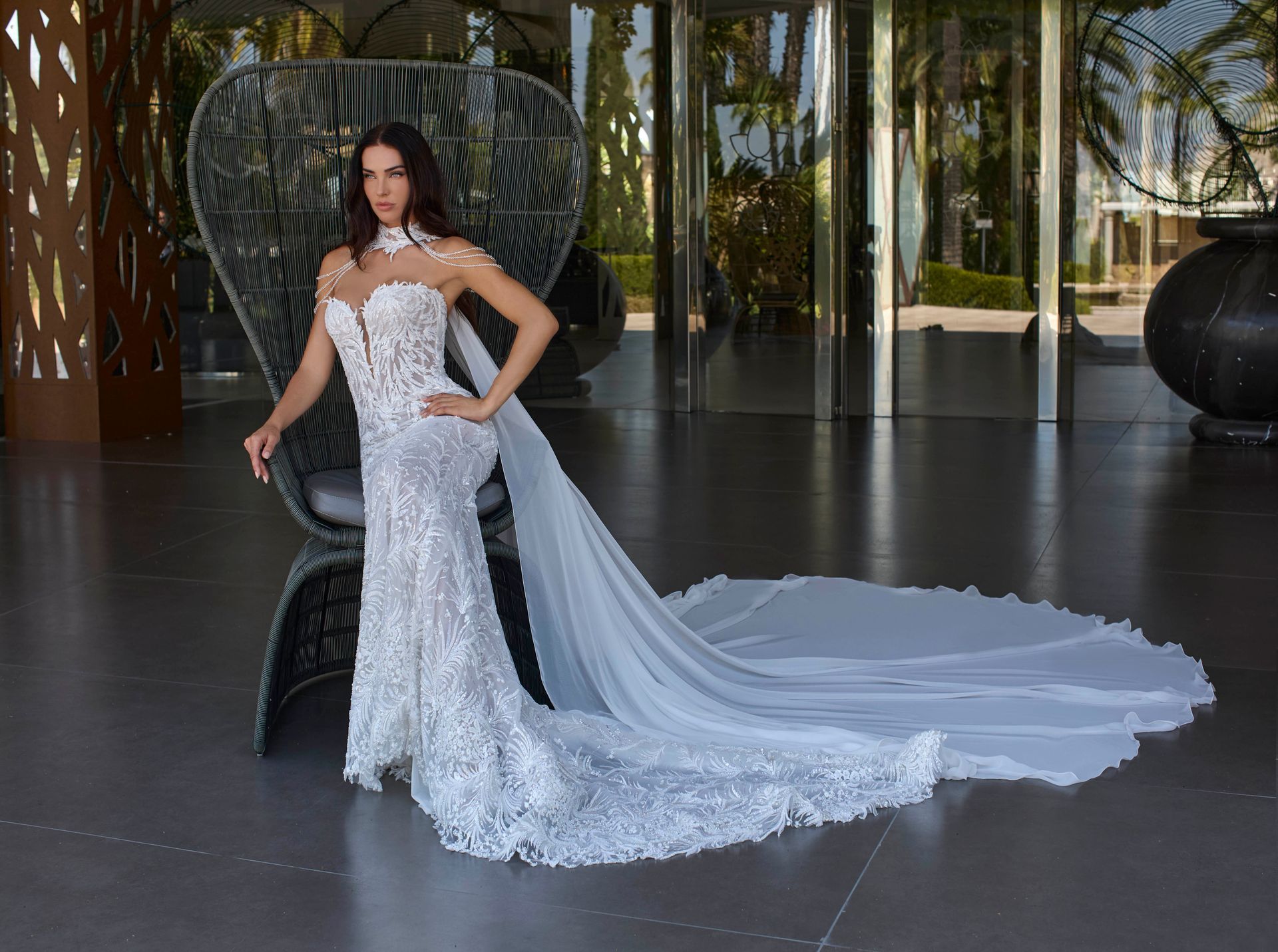 Una donna in abito da sposa bianco decorato con lungo strascico posa vicino a una sedia in un elegante edificio.