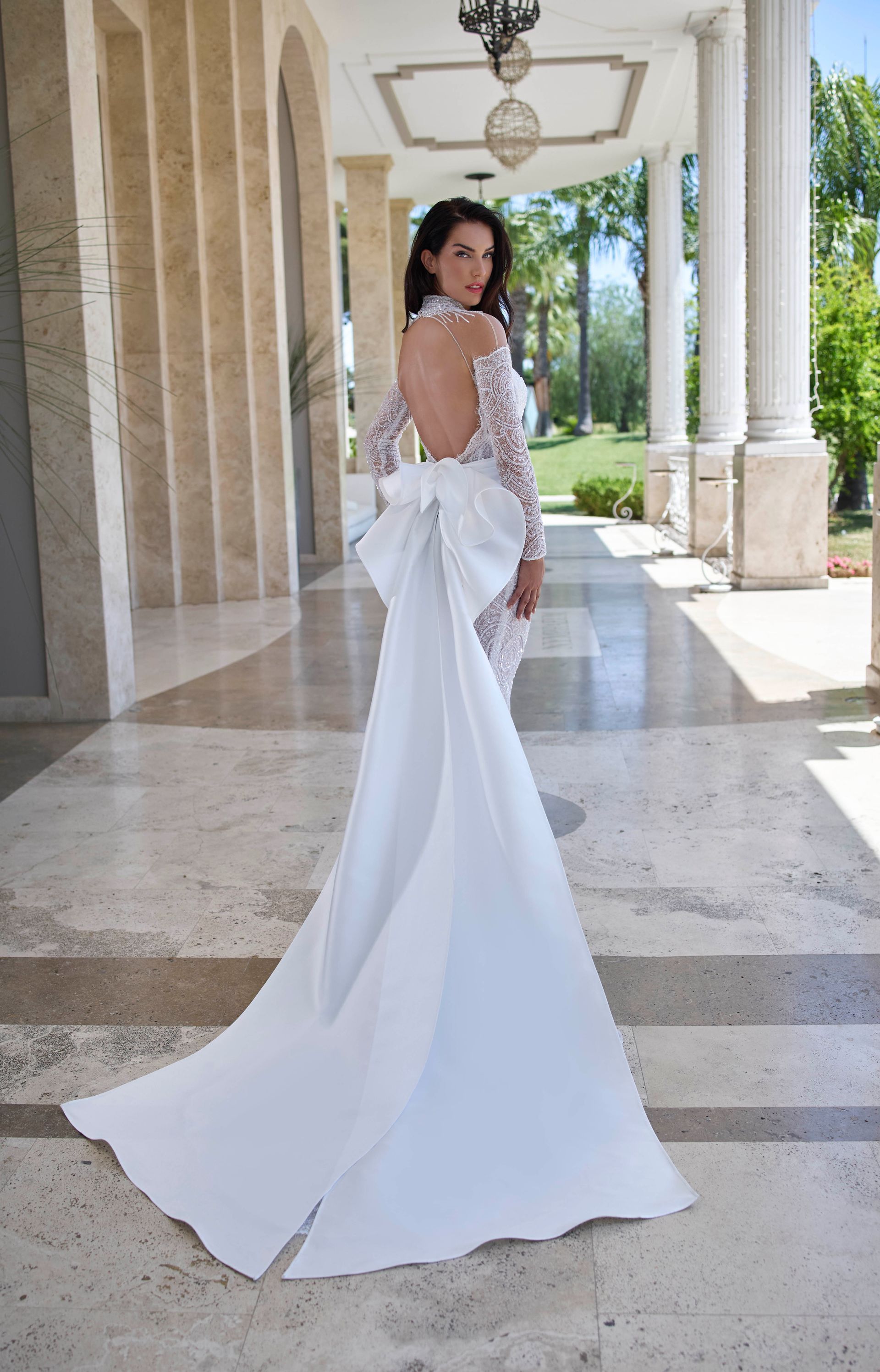 Donna in abito da sposa bianco con lungo strascico e schiena scoperta, in piedi all'aperto.