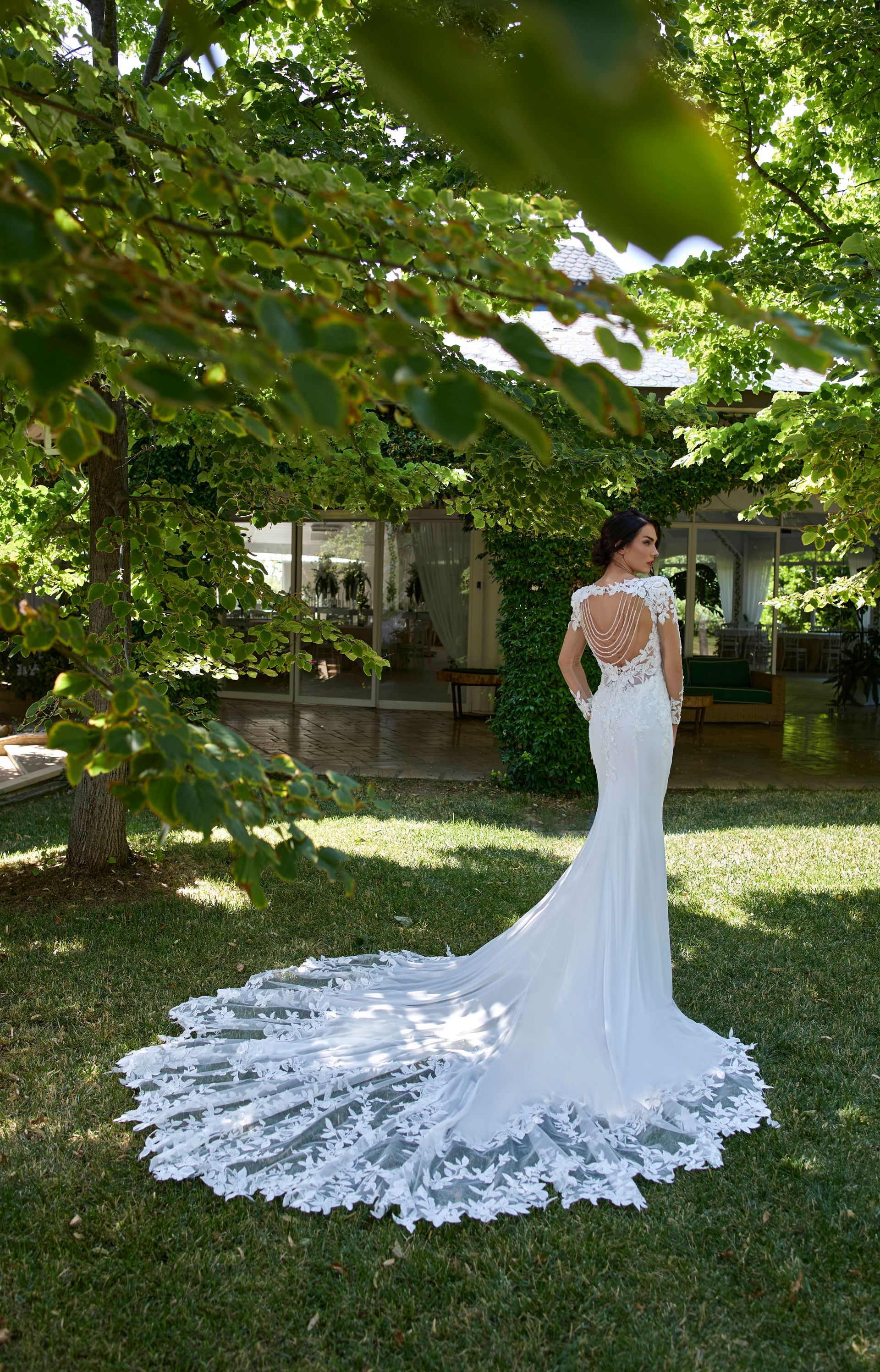 La sposa in abito da sposa in pizzo bianco con lungo strascico posa all'aperto, circondata dagli alberi.