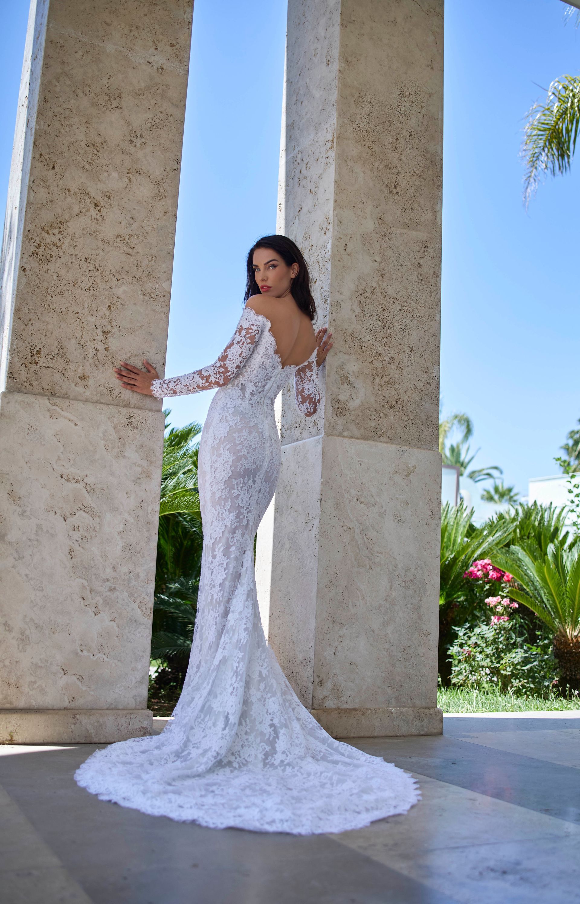 Donna in abito da sposa in pizzo bianco appoggiata a colonne di pietra con un lungo strascico, all'aperto.