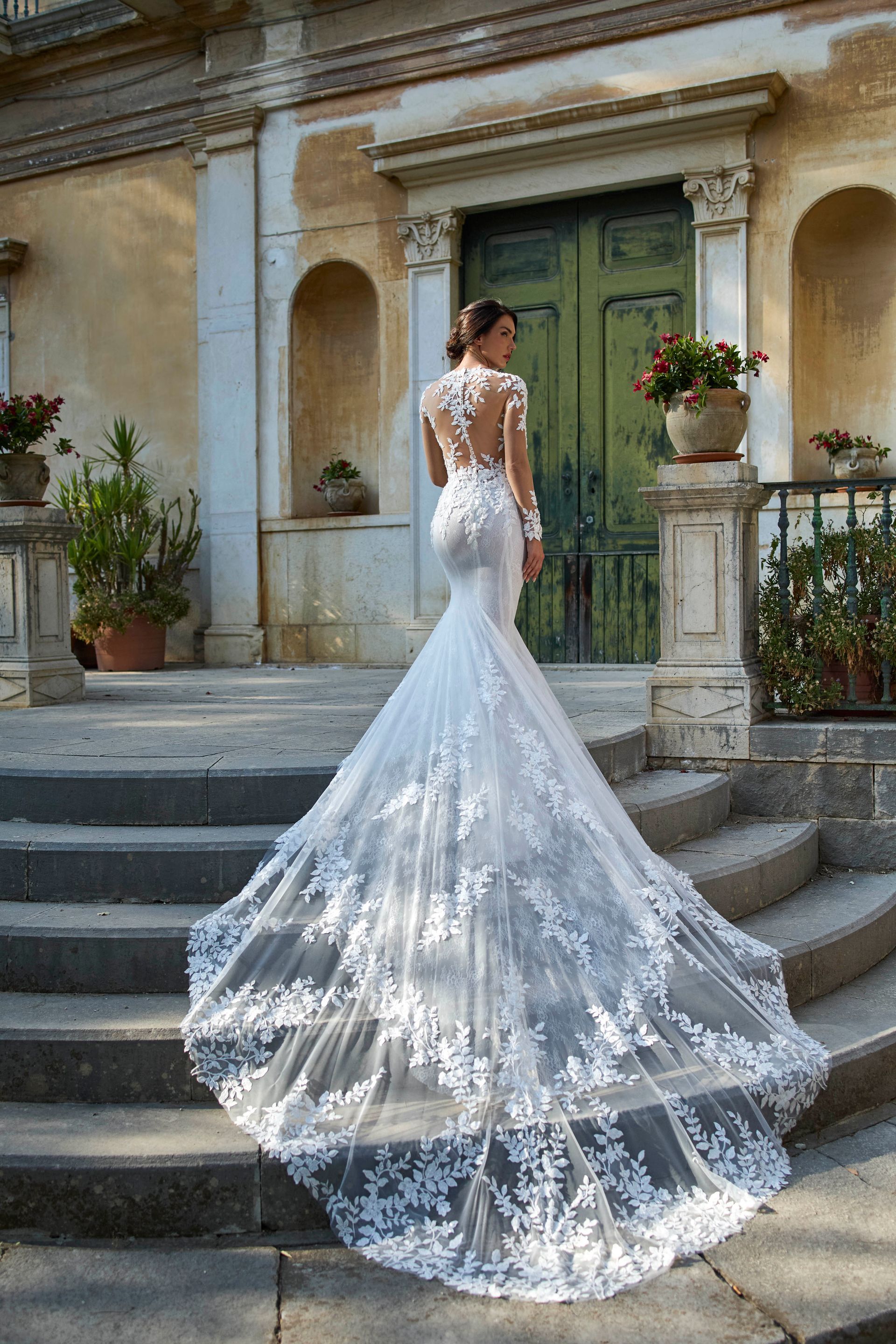 Una donna in un abito da sposa in pizzo bianco con lungo strascico posa sui gradini esterni.