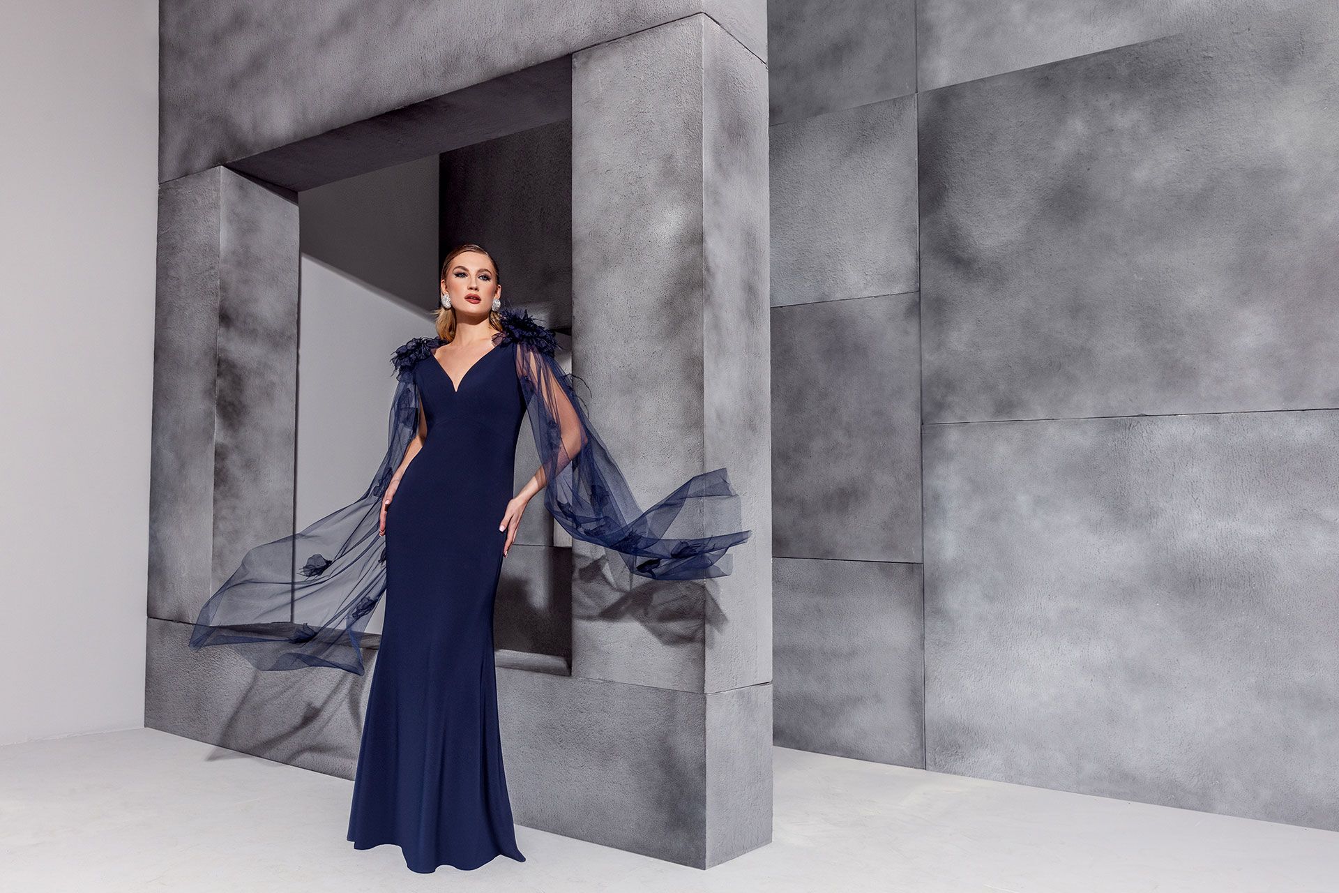 Donna in abito da sera blu navy con maniche svolazzanti, appoggiata a una parete grigia testurizzata.
