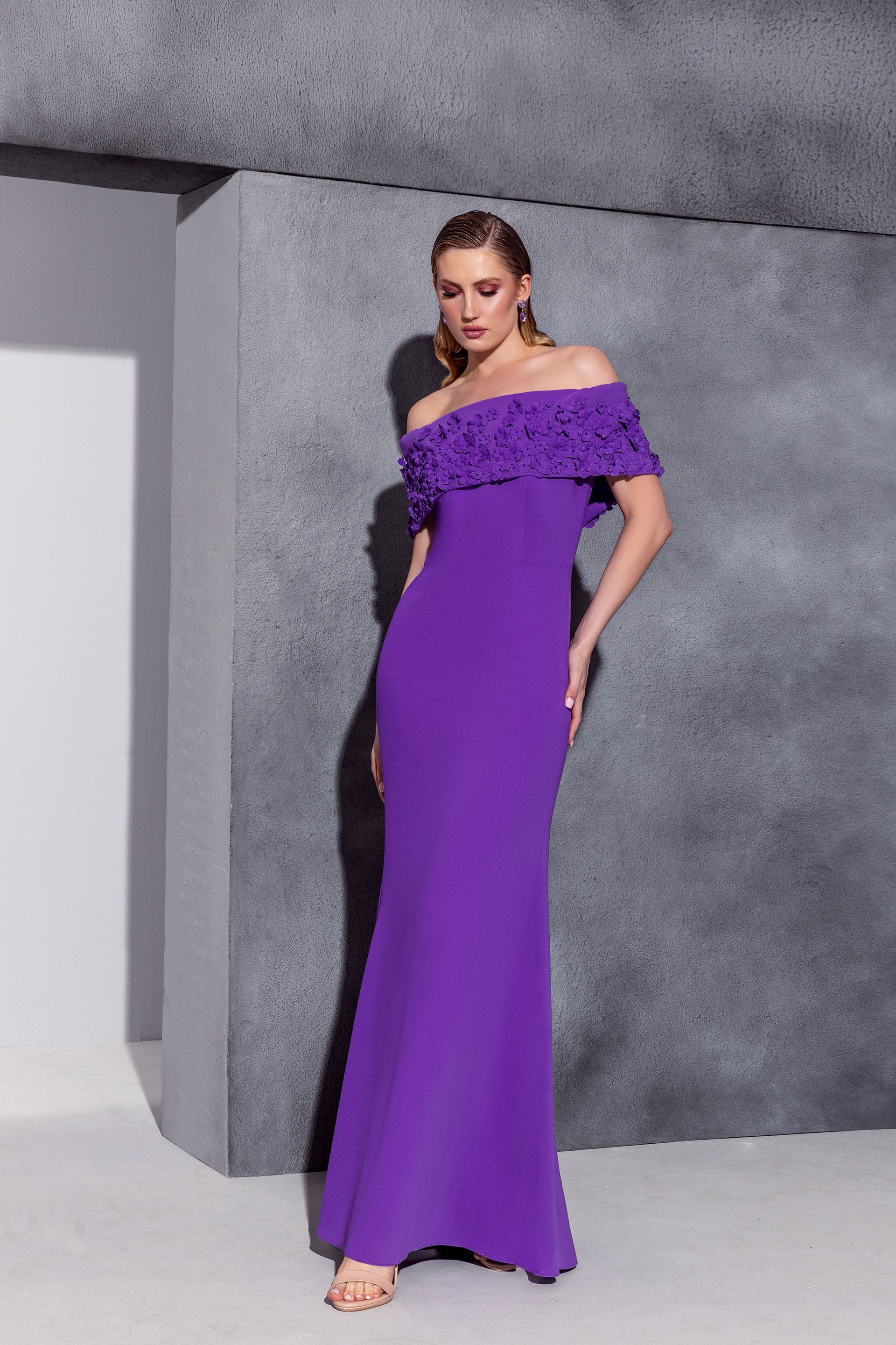Donna in abito viola con spalle scoperte, in piedi davanti a un muro grigio.