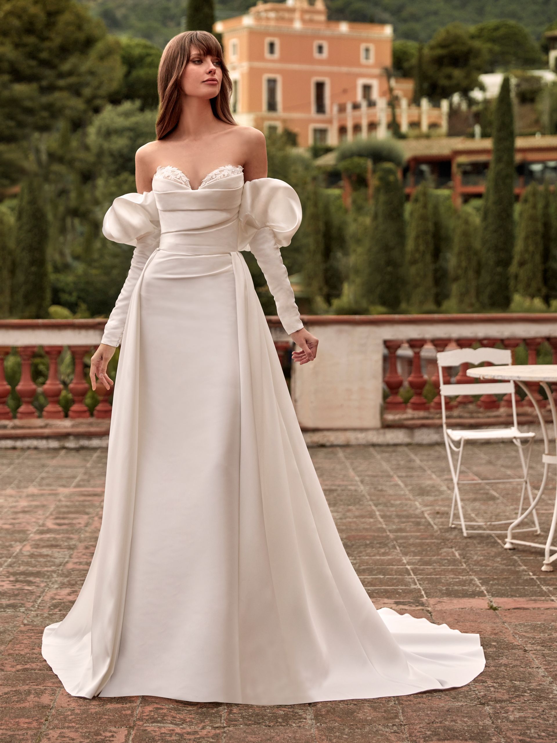 Donna in abito da sposa bianco sporco, con spalle scoperte e maniche staccate, in piedi su un balcone.